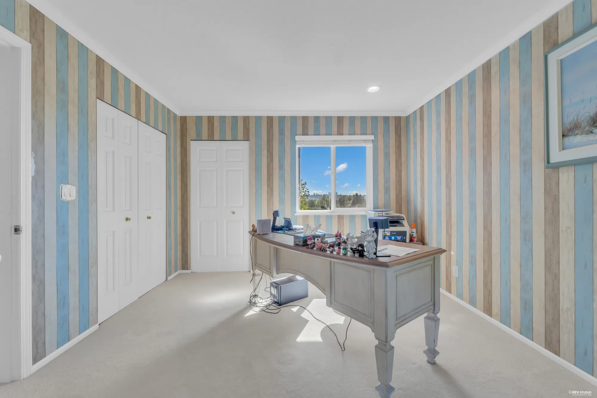 A bedroom with blue striped wallpaper photographed as an office, a desk by the window, closet doors