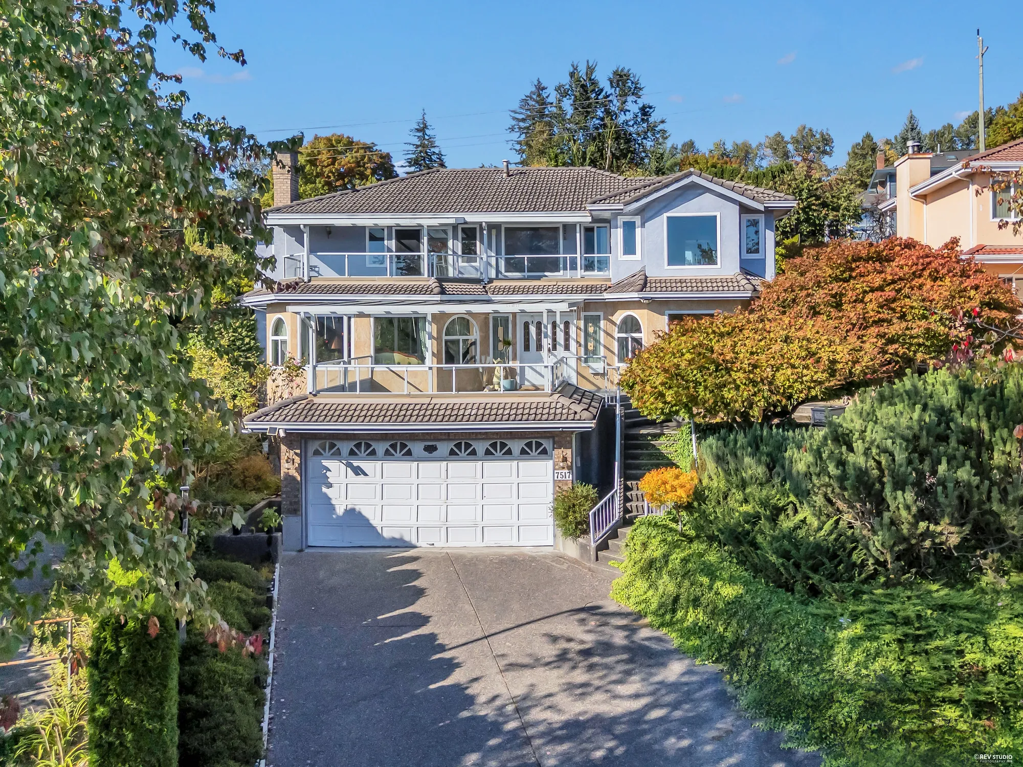 The house from above the driveway in daylight: the double garage, the glass-roofed terrace over it, the balcony along the upper floor, the blue-grey stucco and the trees either side