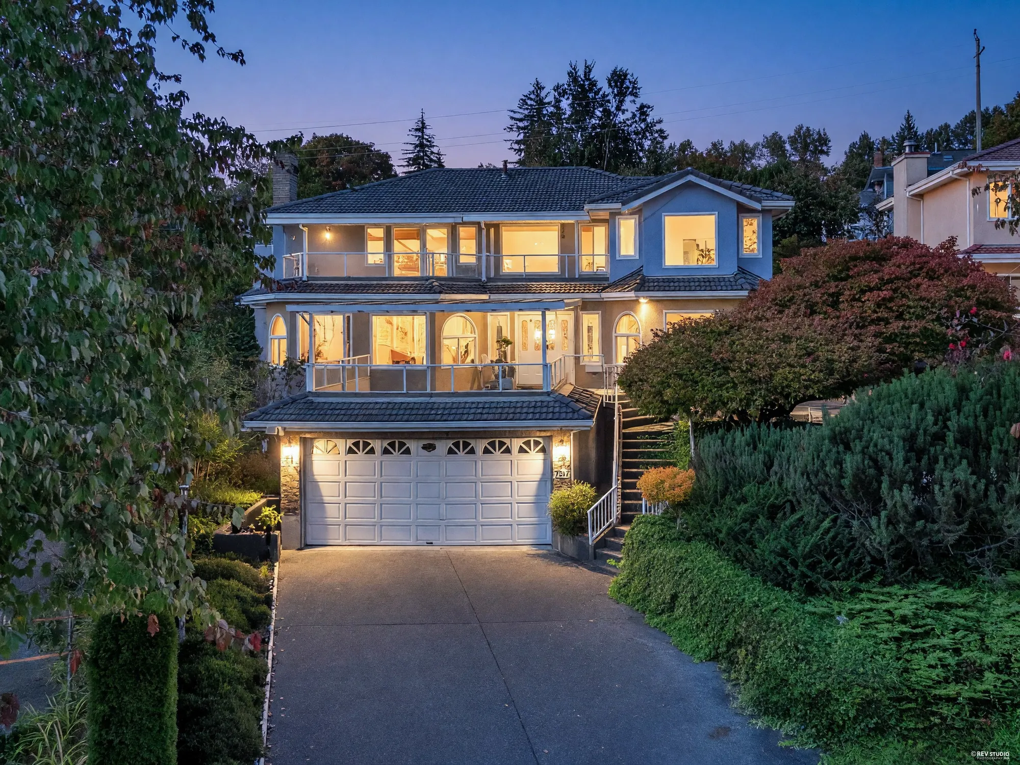 7517 Kilrea Place from above at dusk: the double garage at the driveway, the glass-roofed terrace over it and the balcony above, the blue-grey house lit against the trees