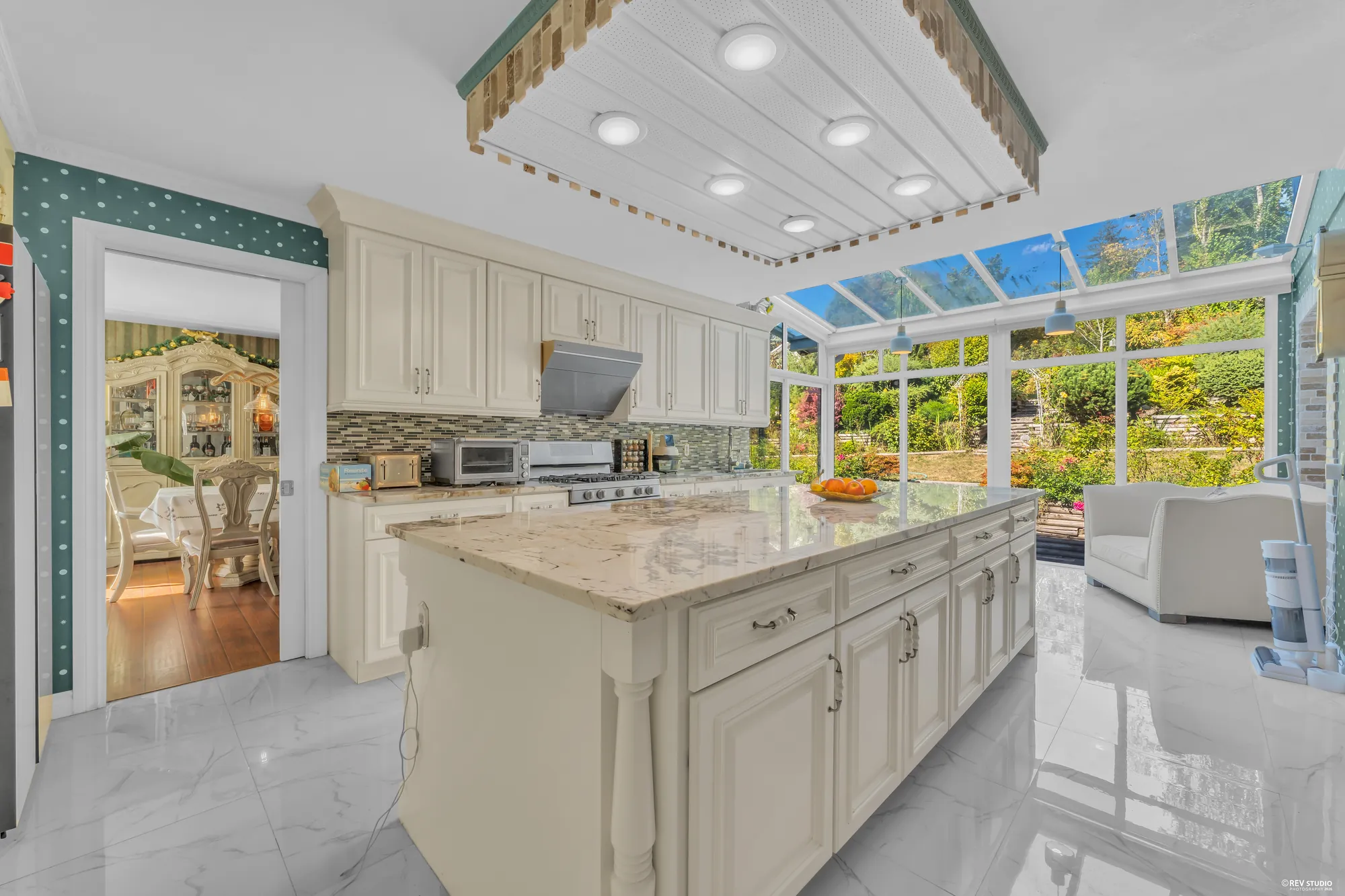 The kitchen: white cabinets, a marble-topped island, the tile floor, and the glass roof of the breakfast nook beyond with the garden through it