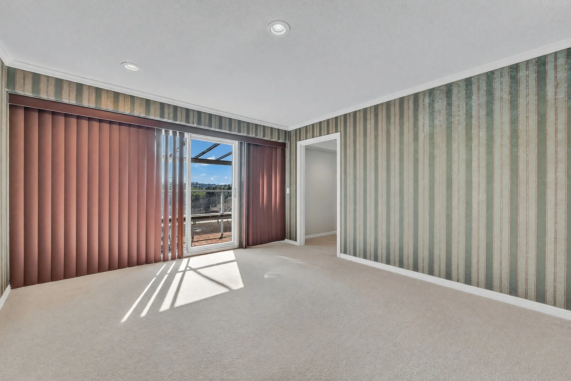 The primary bedroom shown empty: carpet, striped wallpaper, vertical blinds and the door to the balcony, the door to the bathroom