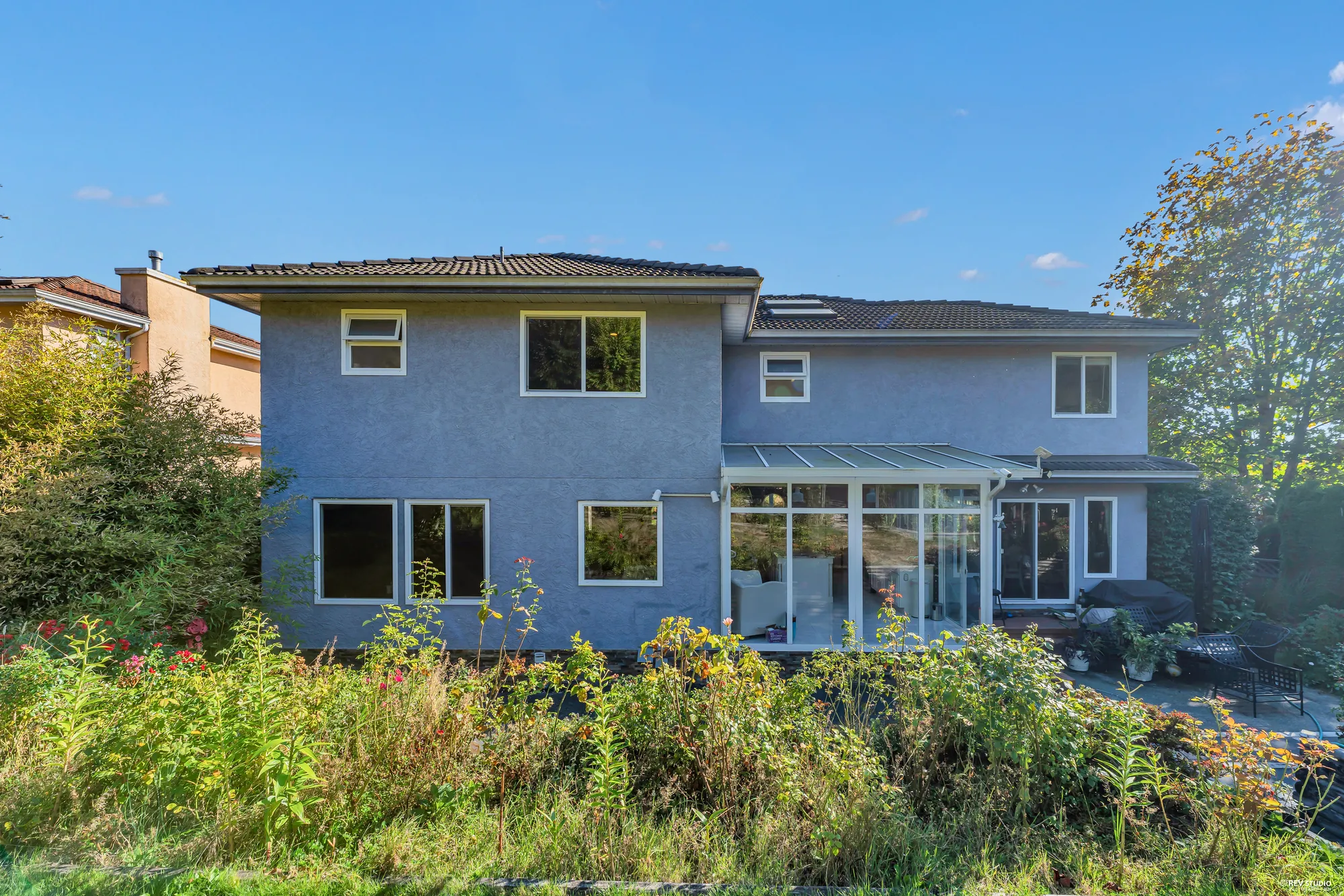 The back of the house from the garden in daylight: blue-grey stucco, the glass sunroom of the kitchen and family room, a patio, the lawn in front